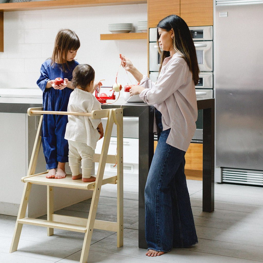 A mother and two daughters using and apple peeler while daughters share standing in a double wide learning tower