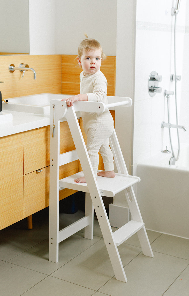 A young girl standing in her white folding learning tower in a bathroom posing white looking at the camera. 