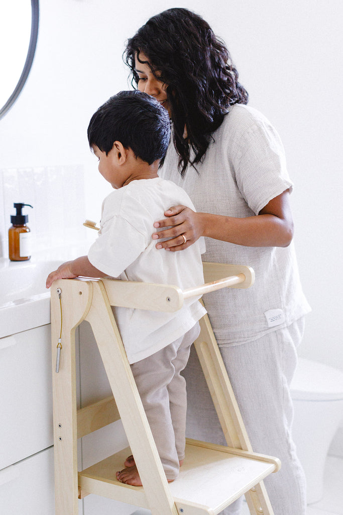 A mom standing beside her son who is in a folding learning tower while they prepare to brush his teeth in a bathroom. 