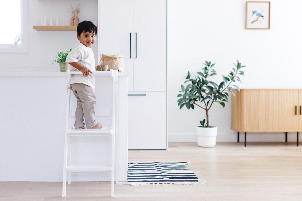 A boy standing up at the counter in his white learning tower and turning backwards to look at the camera. 