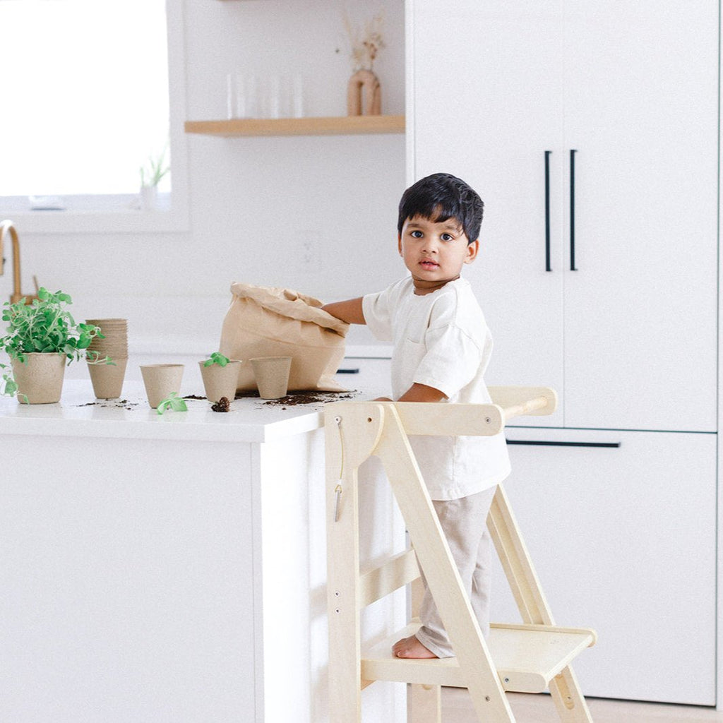 A boy looking back at the camera while playing with dirt and planting seedlings while standing in his learning tower up at the counter. 
