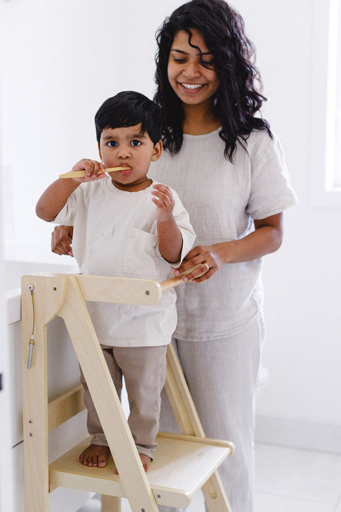 A son brushing his teeth while standing in a learning tower and mom overlooks. 