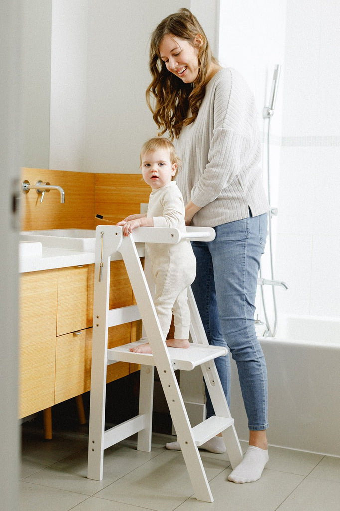 A young girls and mom in the bathroom brushing teeth while girls stands in her white learning tower. 