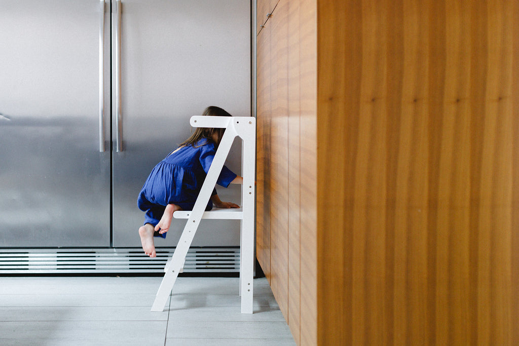 A girl in a royal blue dress climbing up into a white foldable learning tower against the cupboards. 