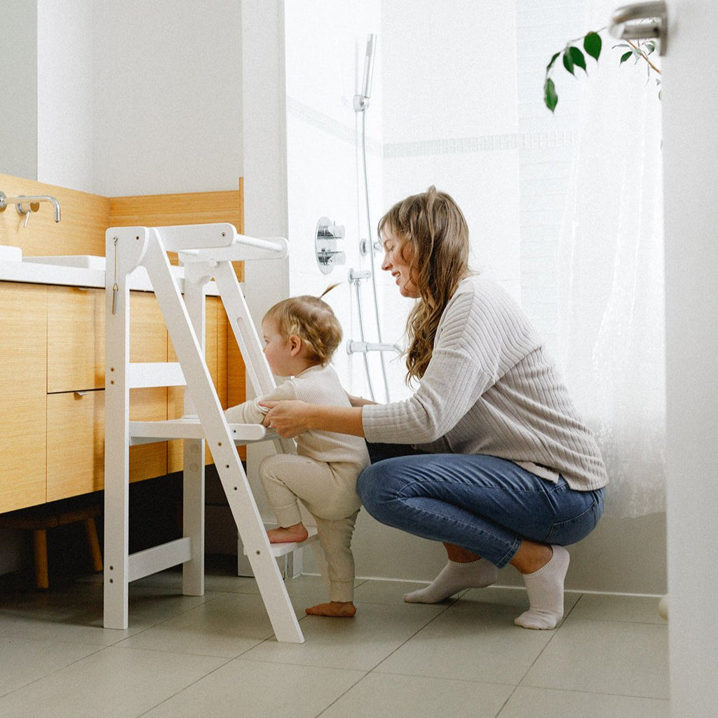 A mom directing her young girl how to climb into the white folding learning tower. 