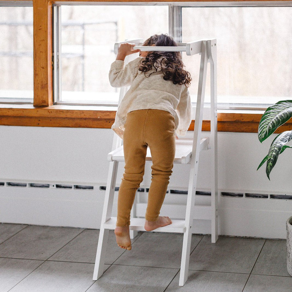 A girl climbing up into her learning tower to draw on the windows. 