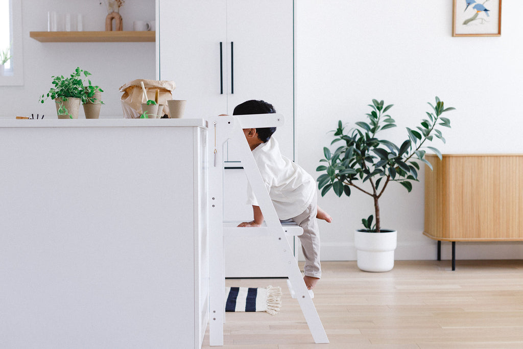 A boy climbing up into his white learning tower to reach the counter height. 