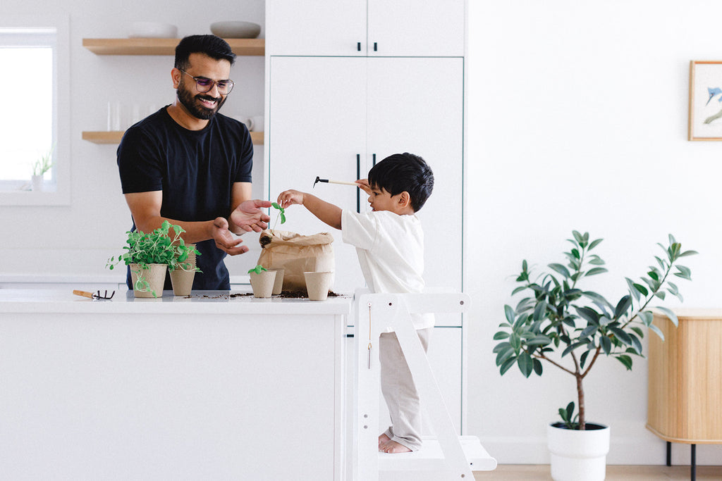 A boy and his dad planting seedlings with soil as the boy stands in his white foldable learning tower. 