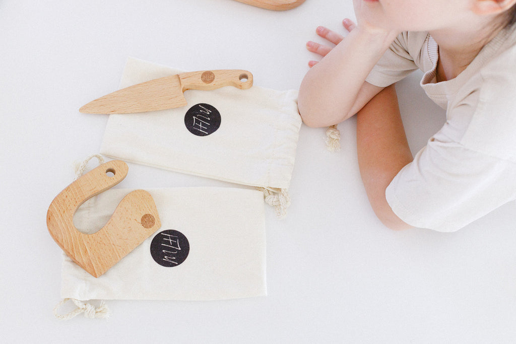 A girl admiring two beginner wooden toddler knives laying down in front of her. 