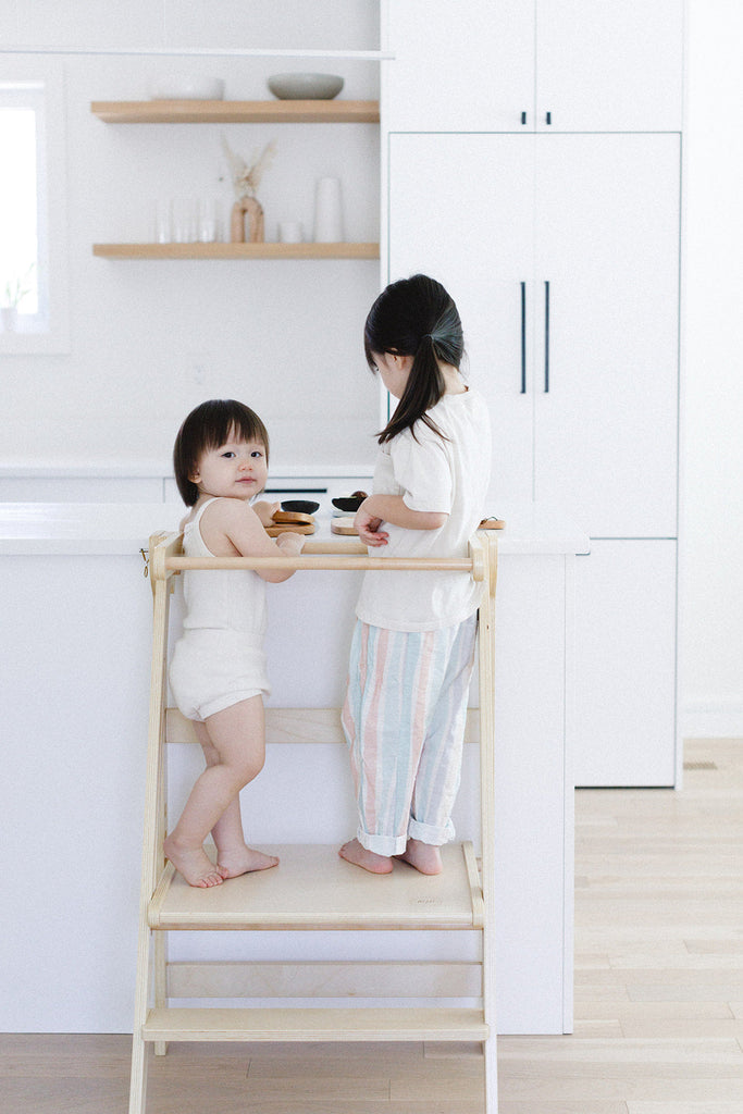 A pair of sisters playing on the kitchen counter while standing in their double wide learning tower