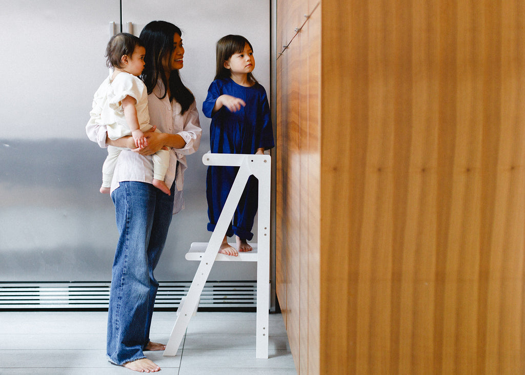 A girl standing in a learning tower looking at cupboard while her mom onlookers with sister in hand. 