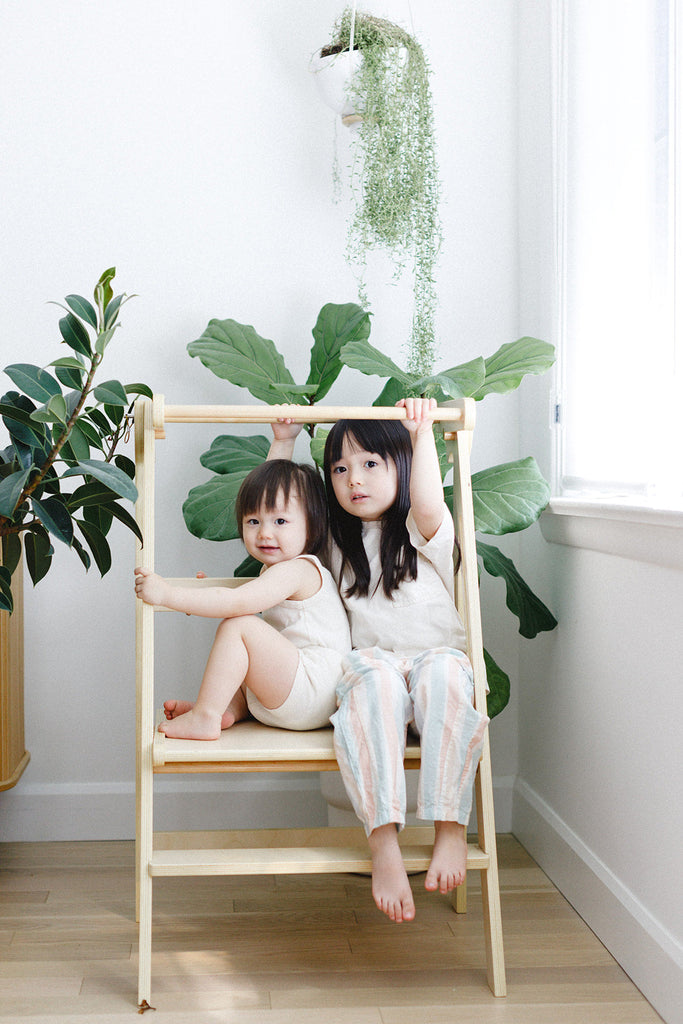 A pair of sisters standing and hanging off a wooden double wide learning tower with plants in the background. 