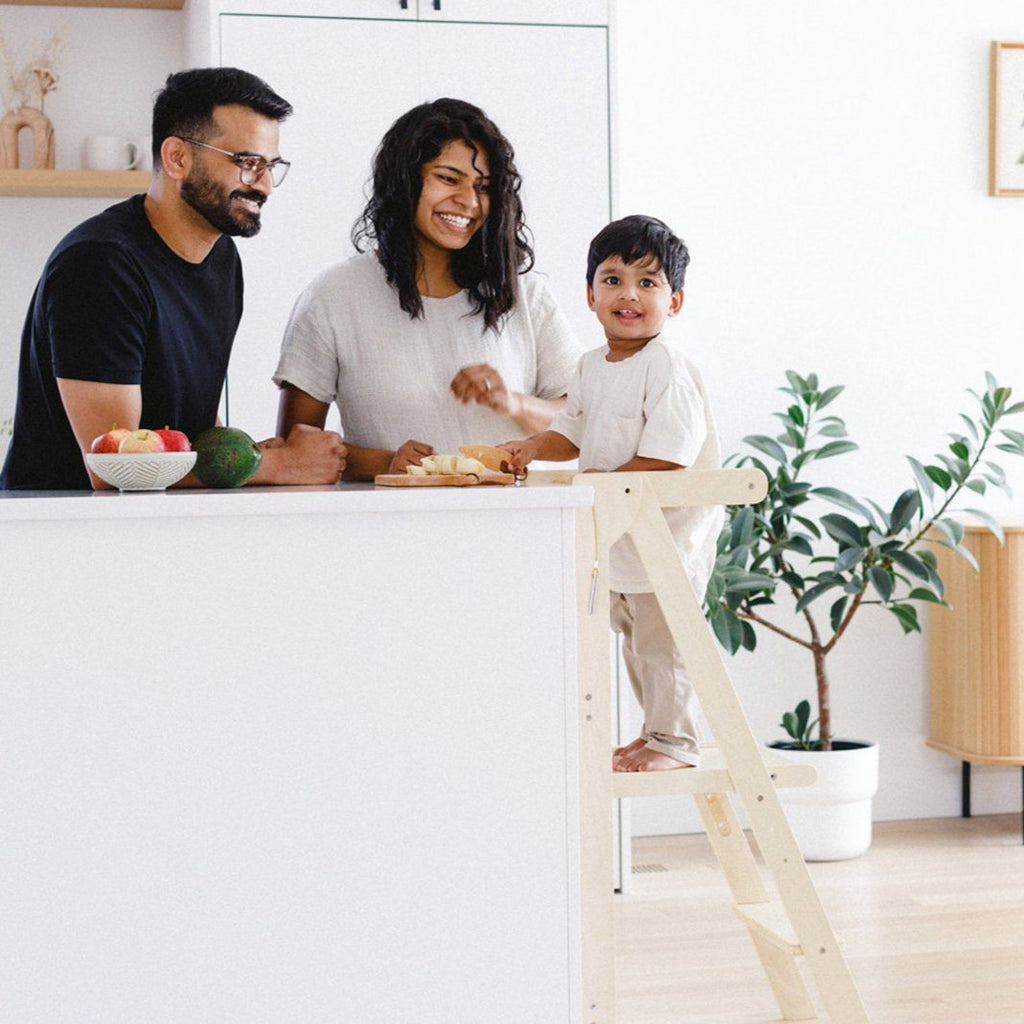 A boy looking at the camera and standing in a collapsible learning tower while his parents look at him with big smiles. 