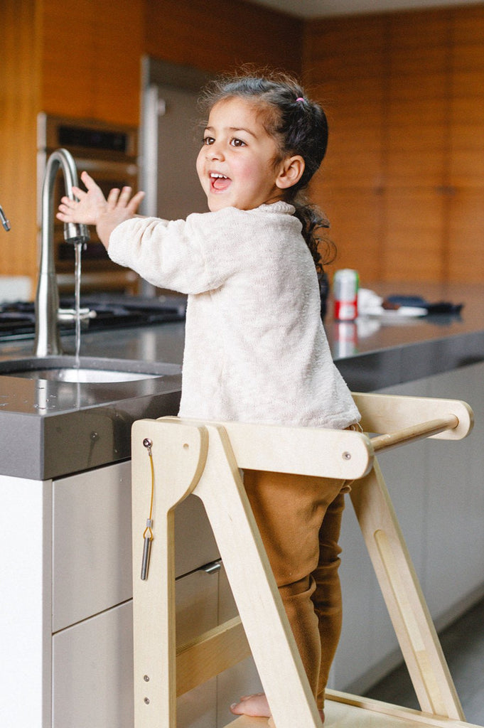 A girls smiles proudly while independently washing her hands using her wooden folding learning tower. 