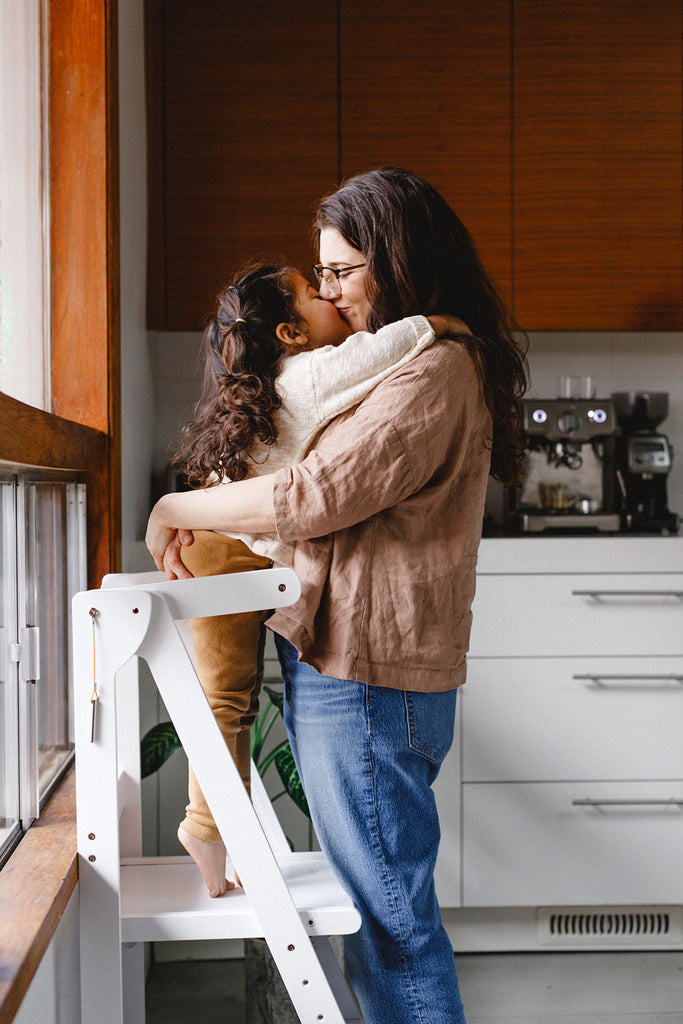 A mom and daughter kissing on the lips while daughter is standing in white learning tower. 