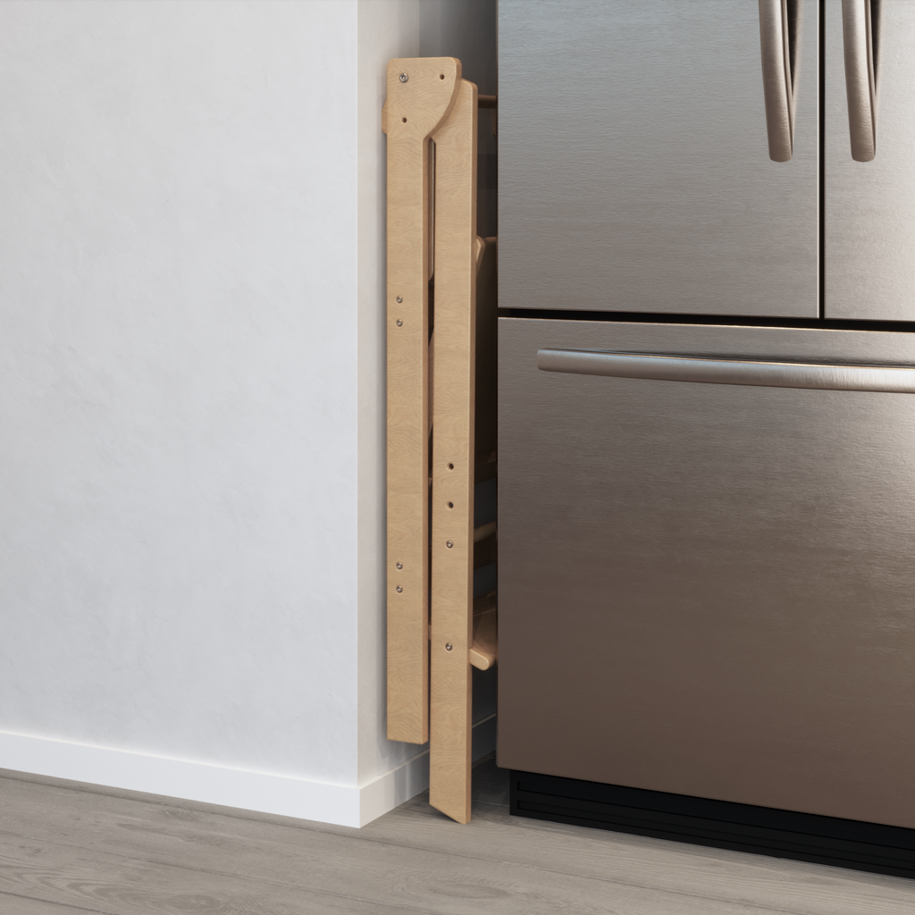 Natural wood learning tower that folds away to store in tight spaces.  The folding learning tower is pictured here tucked away between a fridge and a wall.