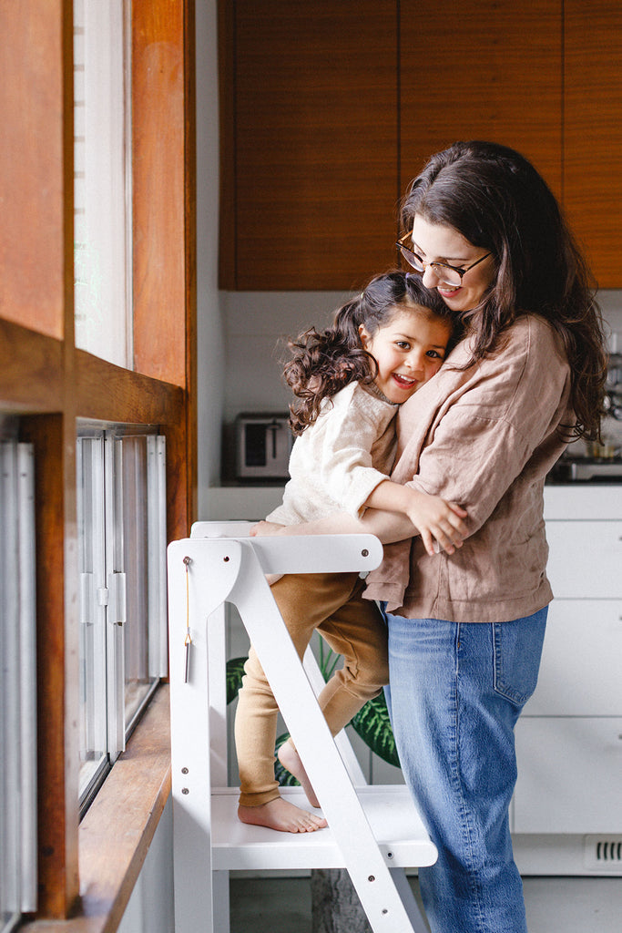A mom and daughter hugging and smiling while daughter is standing in white learning tower and leaning in towards mom. 