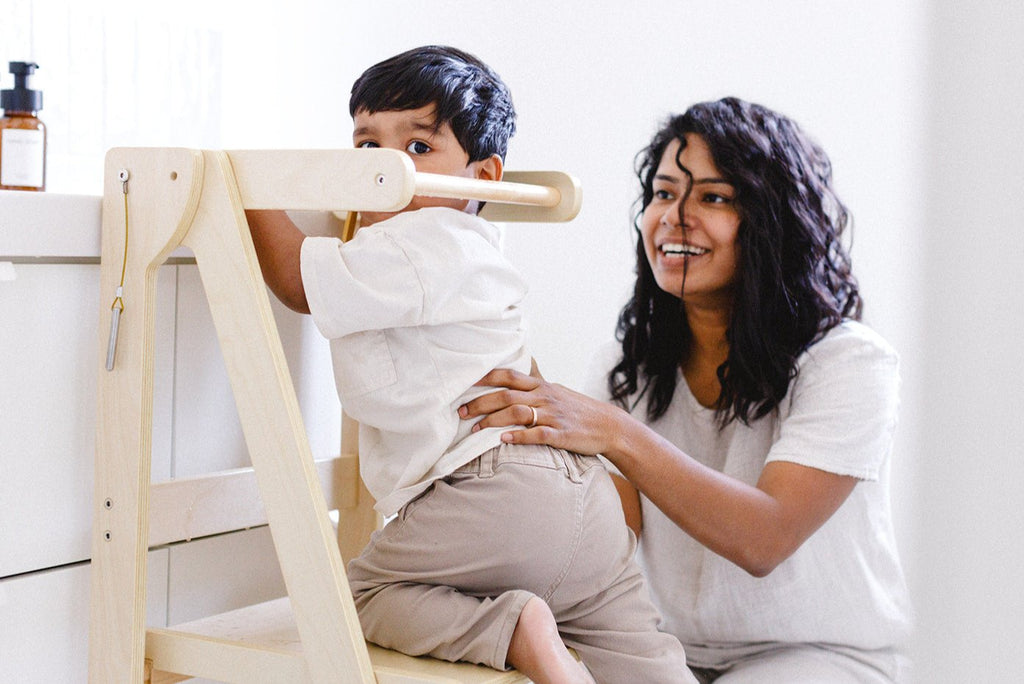 A mom helping her son climb up into his wooden learning tower in the bathroom to wash hands and brush teeth. 