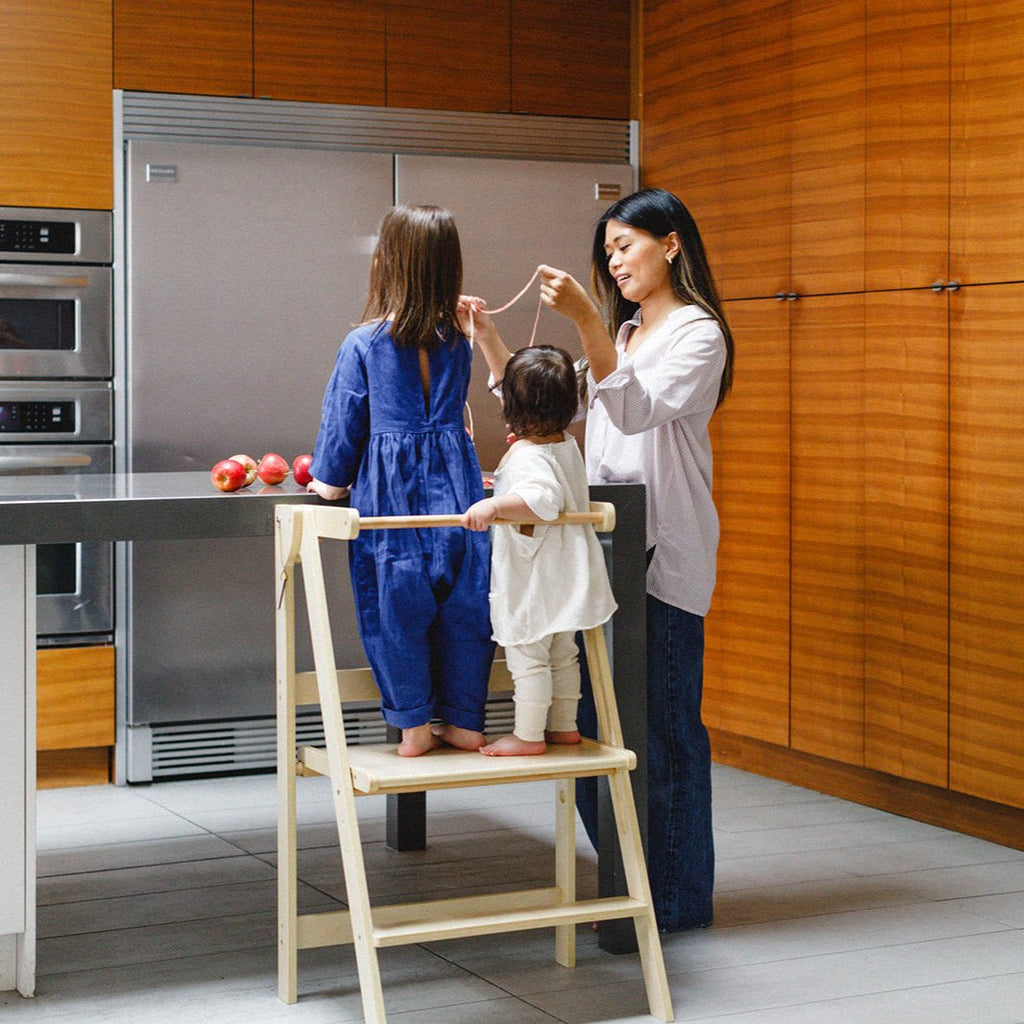 A mom playing with the apple peel after peeling apples while pair of daughters stand in their double wide learning tower. 