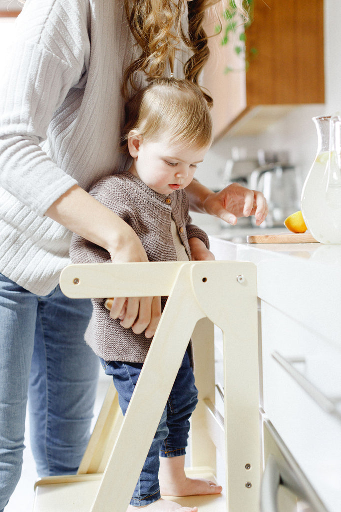 A mom and daughter making lemonade together while daughter is standing proudly in her wooden learning tower. 