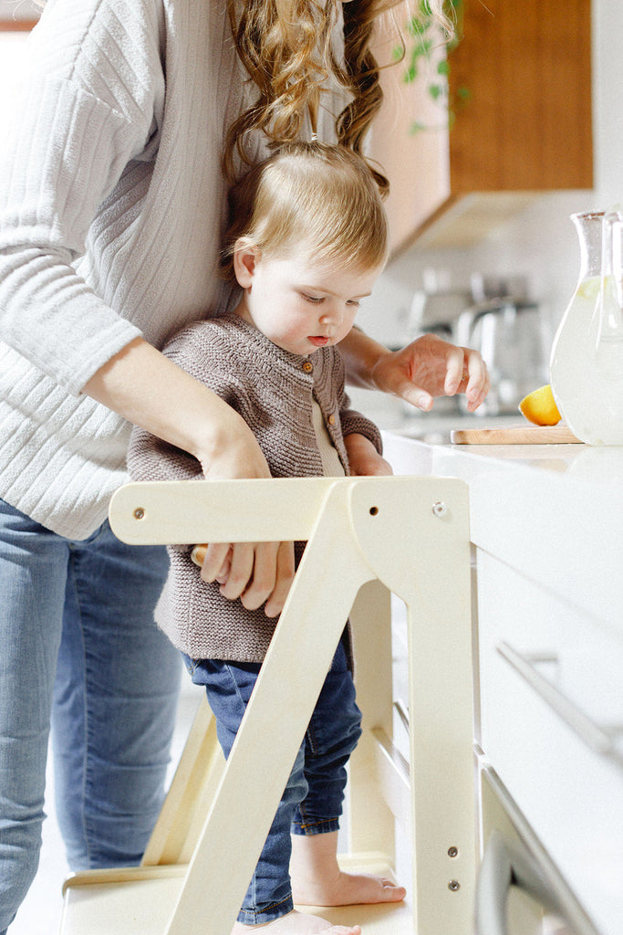 Woman helping a child on a wooden stool in a kitchen.