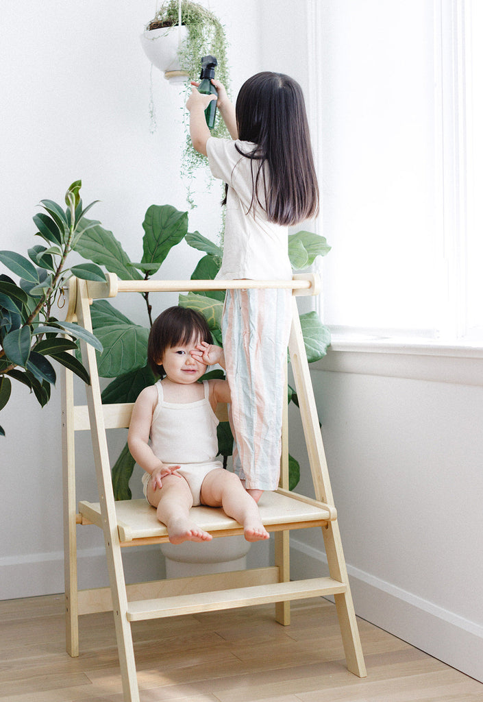 A big sister misting plants while standing in a double wide learning tower with little sis sitting on the bottom