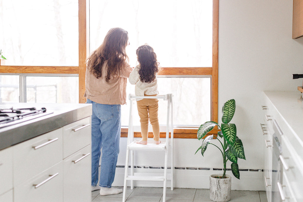 A mom and daughter drawing on the window while the daughter stands in her learning tower. 