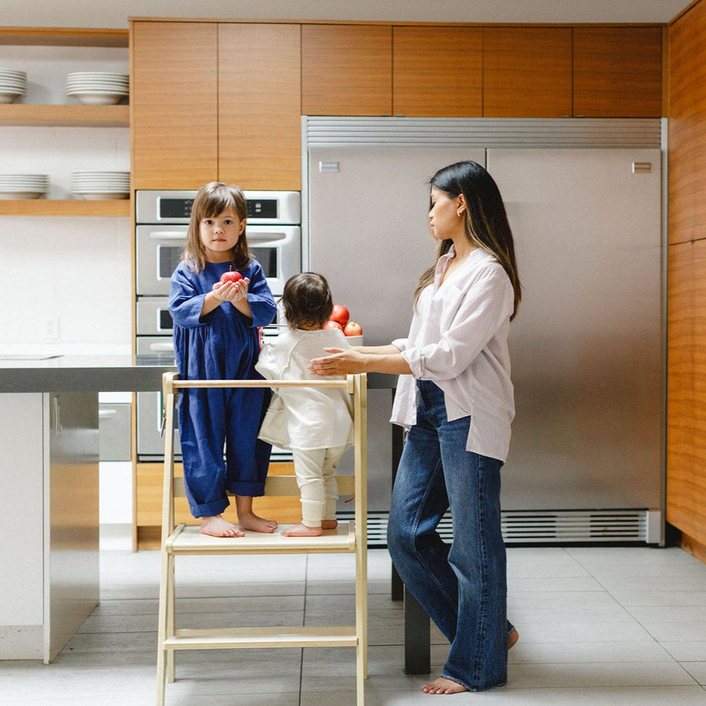 A daughter holding an apple in her hands while sharing the double wide learning tower with her sister and mom onlooking. 