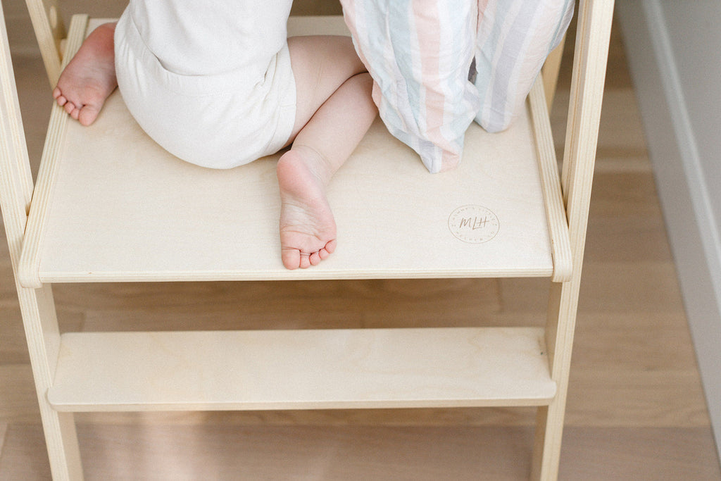 A detailed close up of a pair of sisters feet standing on the wooden platform of a double wide learning tower. 