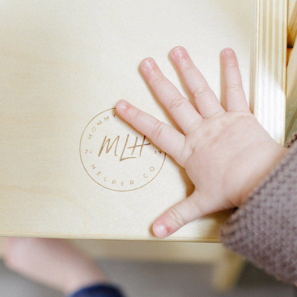 A close up of a little hand touching the platform of a learning tower with the brand logo engraved in the Baltic Birch wood. 