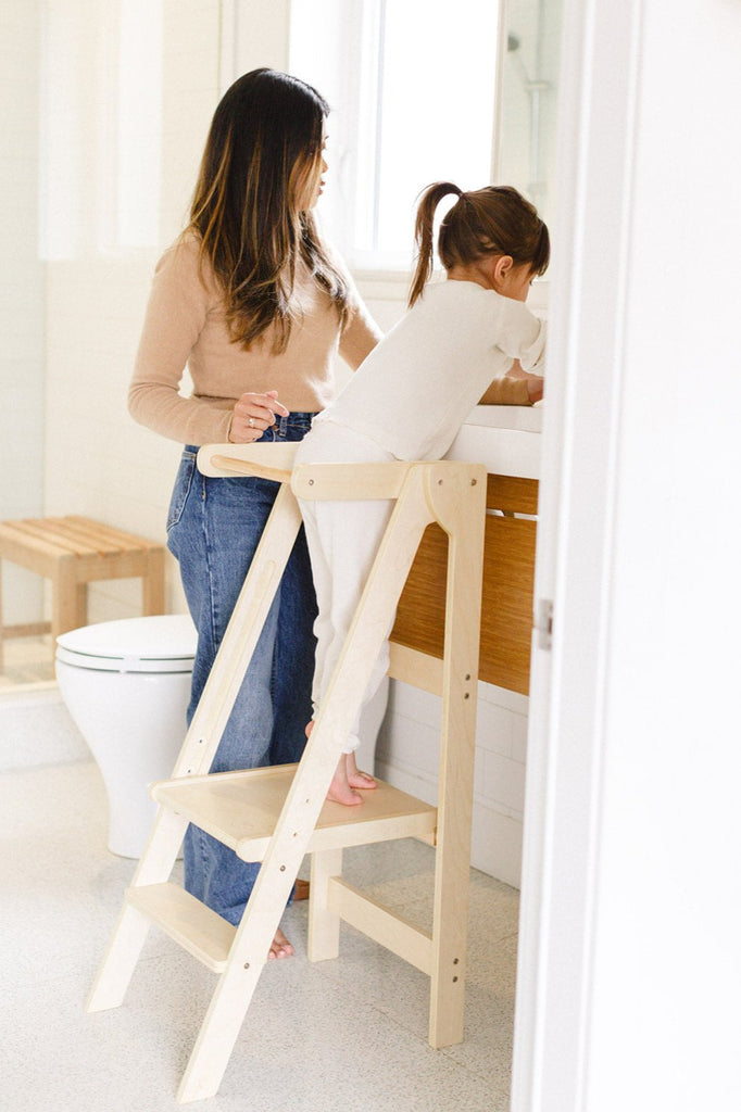 A girl washing hands while standing in a learning tower beside mom. 