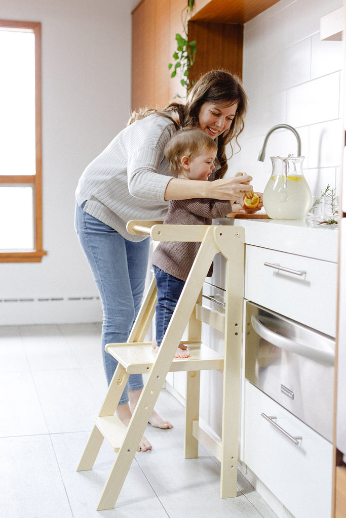 A mom helping her daughter to squeeze a lemon while making fresh lemonade in a learning tower. 