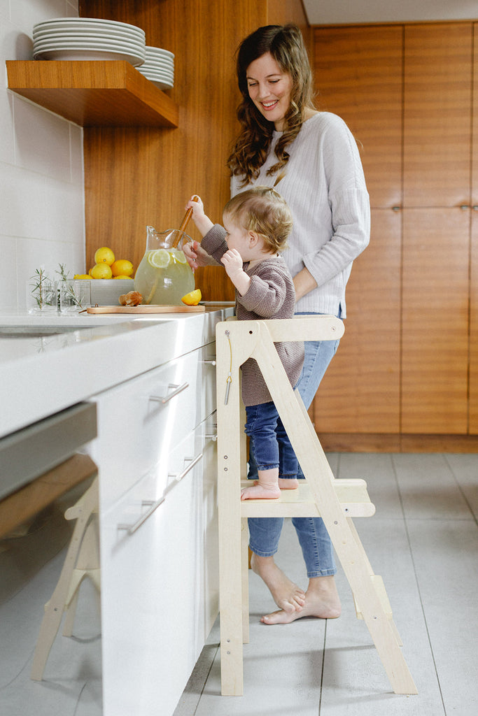 A mom watching while her daughter stirs the lemonade while standing in a wooden learning tower. 