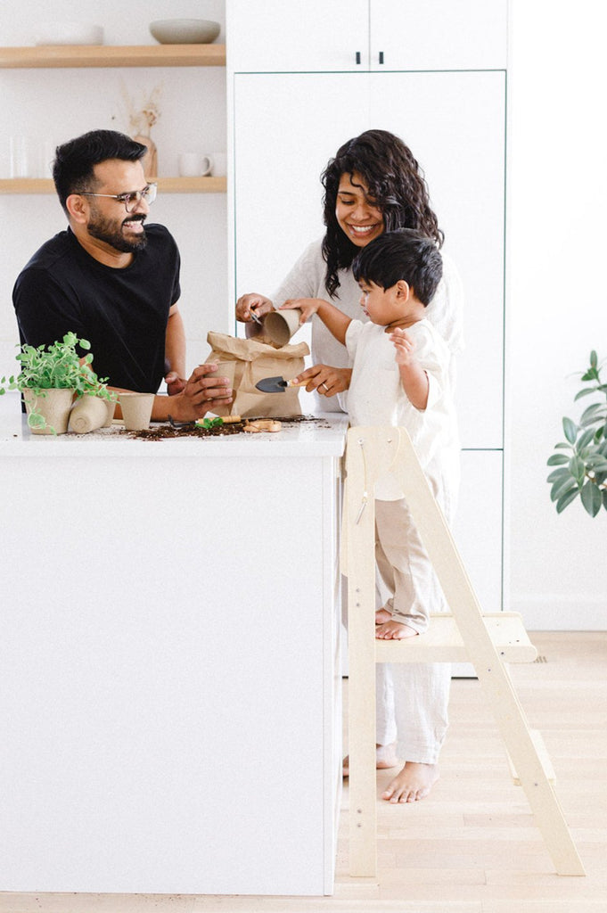 A boy standing in a wooden folding learning tower planting seedlings with his parents onlooking and everyone smiling and laughing. 