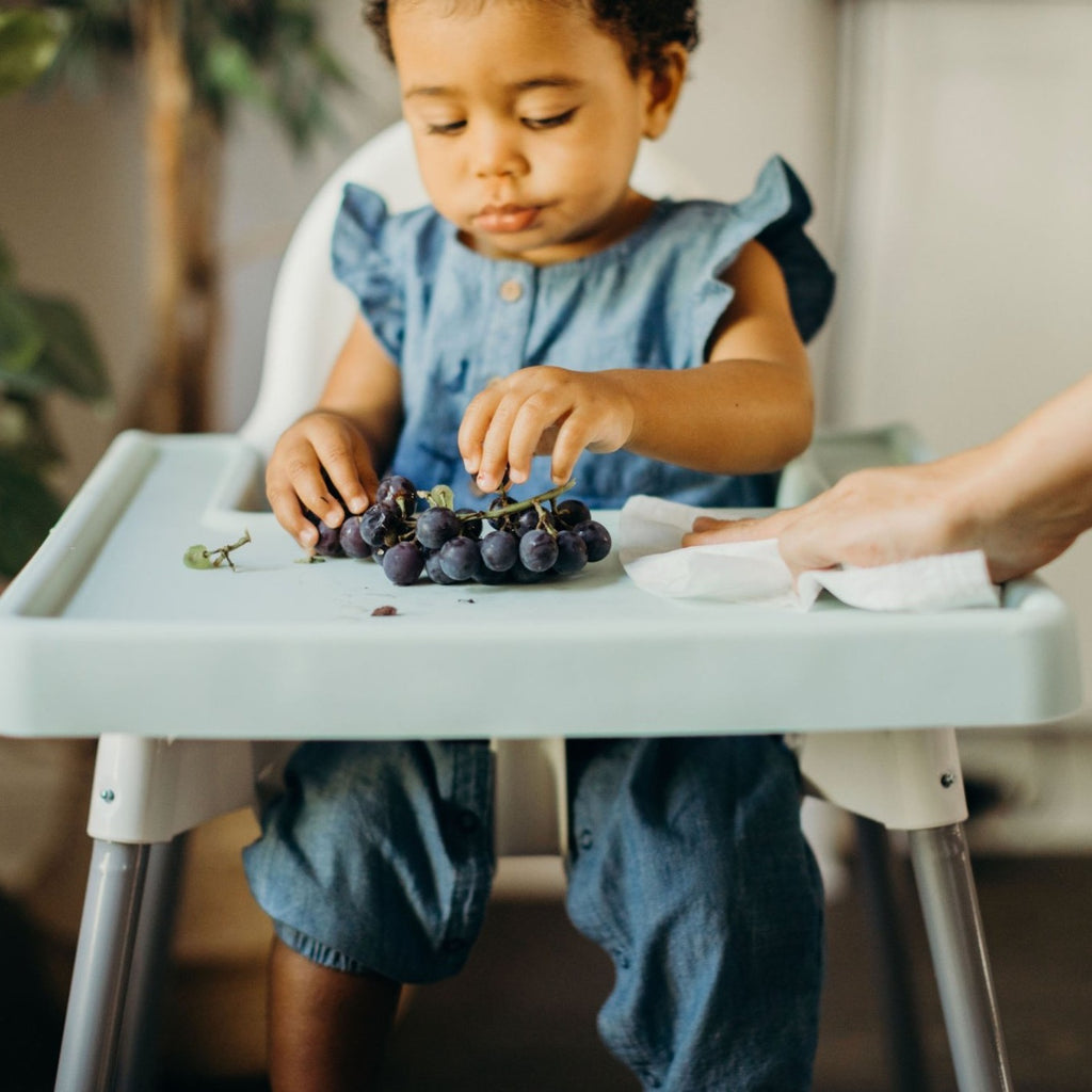 A child eats grapes from the Ikea antilop highchair full coverage placemat cover in mint green while an adult easily wipes the cover down. 