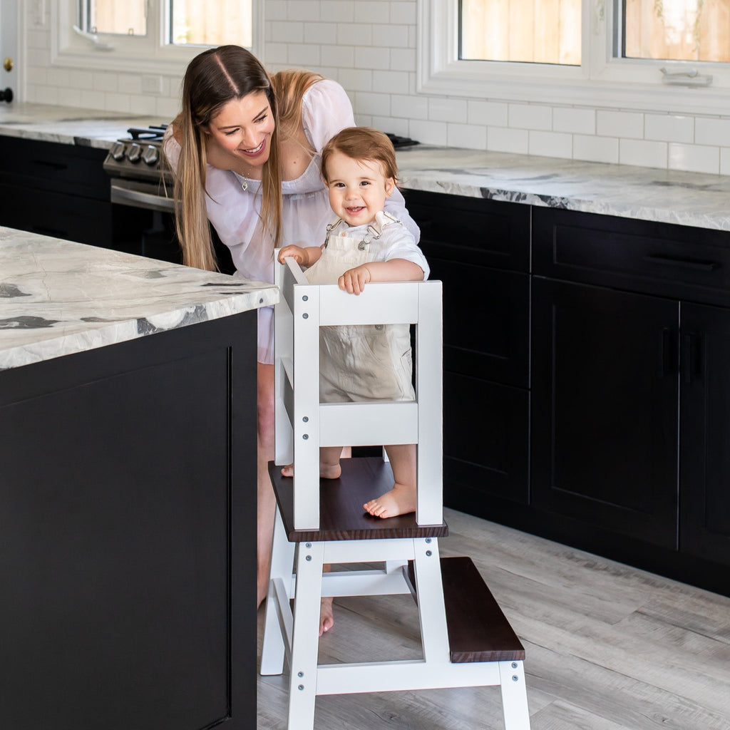 White and Walnut color Classic Montessori Toddler Learning Tower against a kitchen counter - child standing in tower with Mom smiling