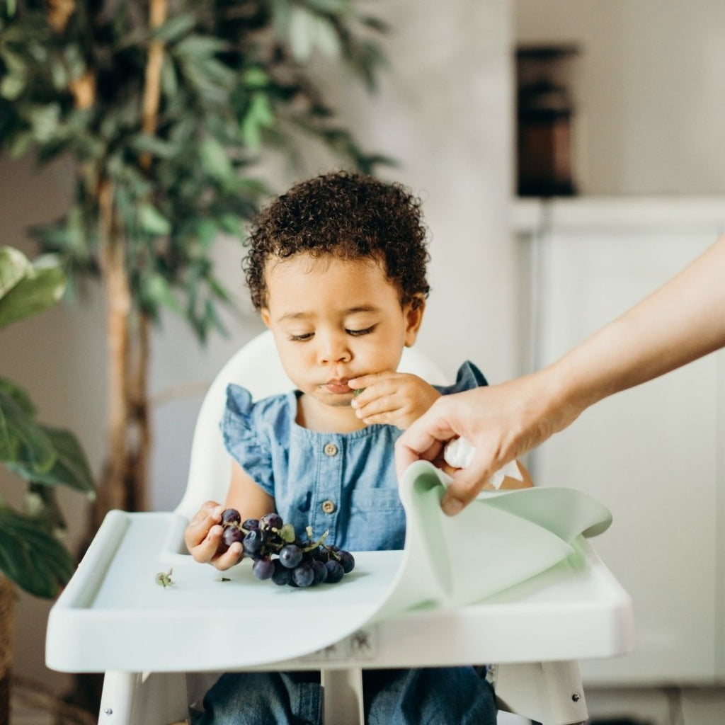 Child sitting in Ikea Antilop highchair eating grapes while parent shows removable full coverage placemate cover