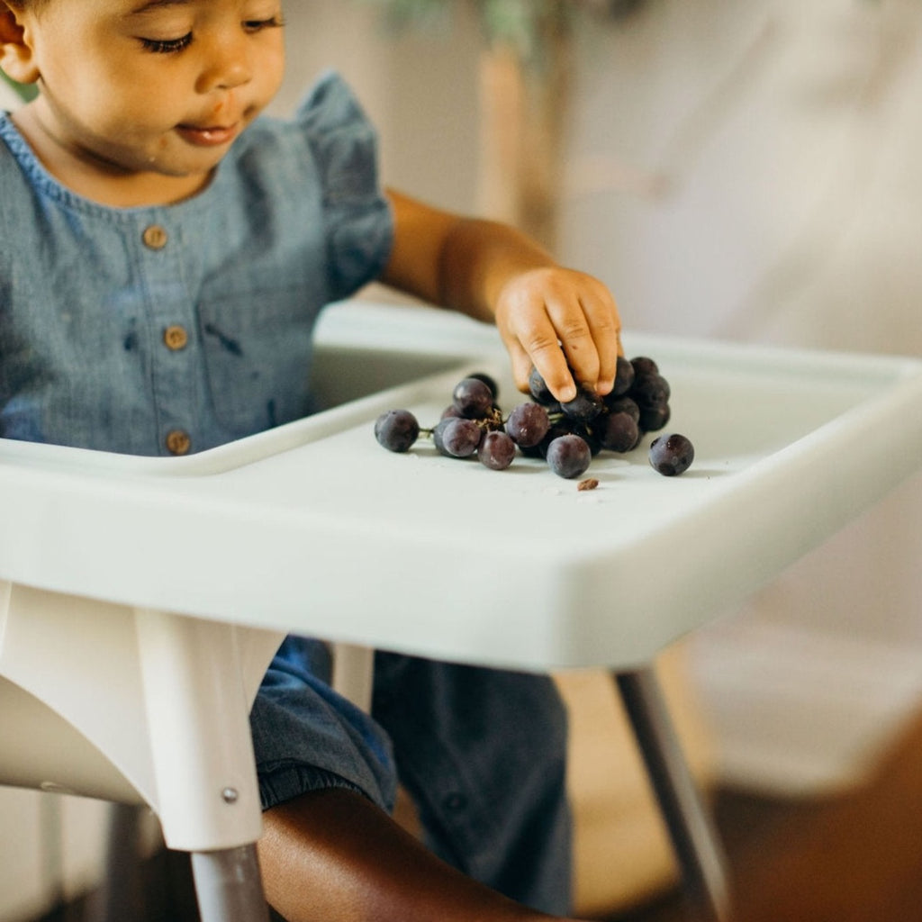 Toddler eating grapes in Ikea Antilop highchair with full coverage placemat cover on top. 