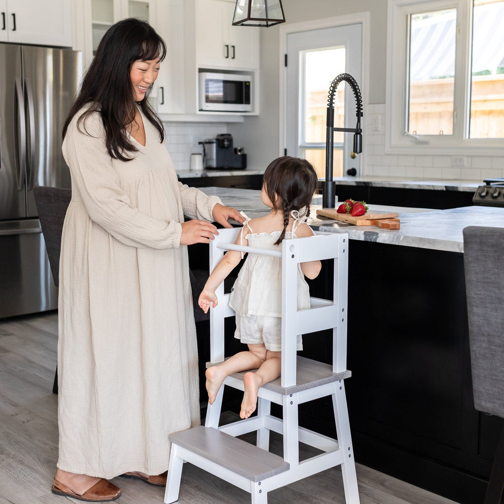 a toddler climbs up and into her learning tower while smiling at mom and wanting to help in the kitchen