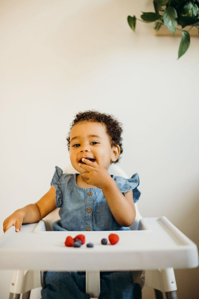 Toddler eating berries while sitting in Ikea Antilop Highchair that has mellow grey full coverage placemat cover on top. 