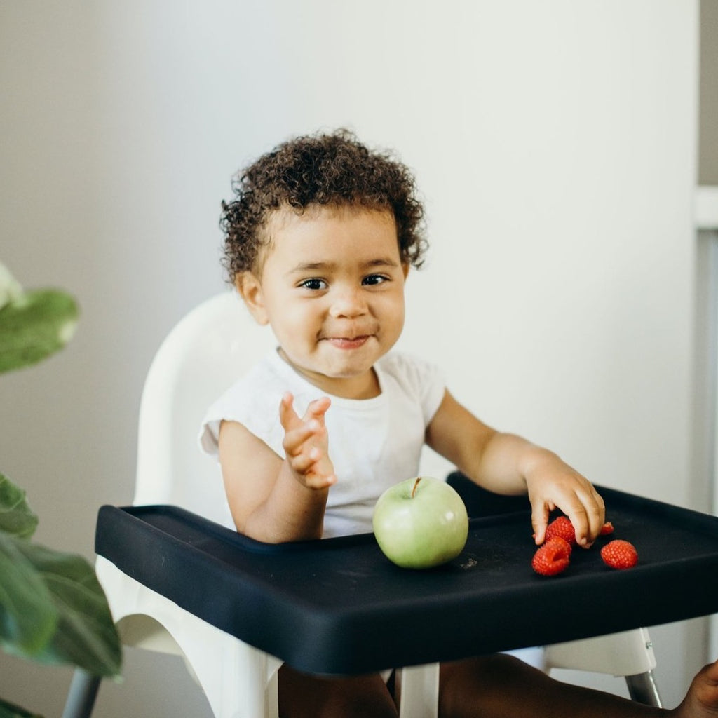Smiling Toddler sitting in Ikea Antilop Highchair eating fruit off of a Charcoal Black color full coverage placemat cover